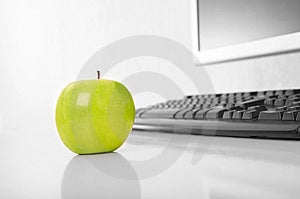 Zelené jablko je na stole, na pozadí klávesnice a monitora.